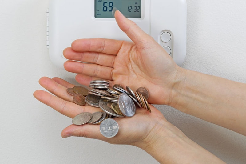 5 Ways to Save Money on Your AC and Summer Energy Bill. Photo of someone holding a handful of coins in front of a digital thermostat mounted on a wall.