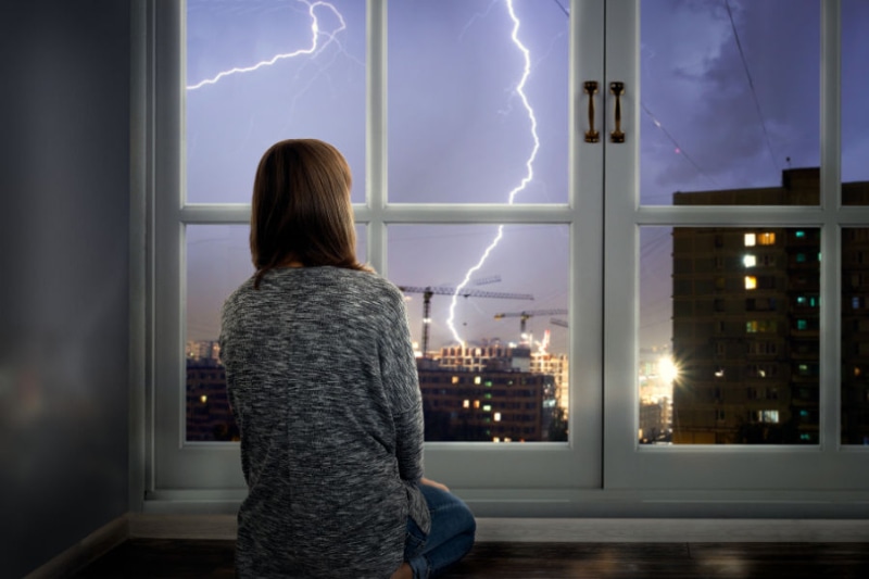 How to Protect Your AC From Storms - Girl looks out the window at a thunderstorm.